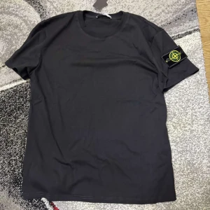 Svart t-shirt från Stone Island - Säljer en svart t-shirt från Stone Island med klassisk rund hals och korta ärmar. Ikonisk logotyp-patch på vänster ärm. Perfekt för en clean och stilren look.