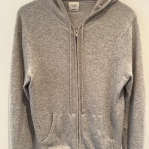 Grå cashmere hoodie från John Henric - Mjuk och lyxig grå hoodie i 100% cashmere från John Henric. Tröjan har dragkedja framtill, huva och två fickor. Perfekt för dig som gillar en clean och chill stil med premiumkänsla. Det är tyvärr två små hål på ställen som ej syns när man har på sig den och där av priset, nypris på tröjan är 2 499kr