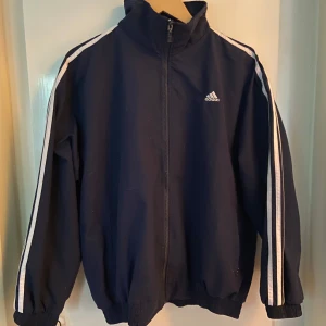 Mörkblå Adidas vindjacka XL - Säljer en mörkblå Adidas vindjacka i storlek XL med klassiska vita ränder längs ärmarna och Adidas-logga på bröstet. Jackan har dragkedja framtill, hög krage och ribbade muddar. Perfekt för dig som gillar sportig och clean stil.