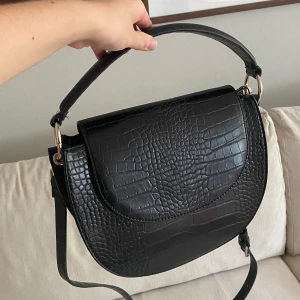 Svart axelväska  - Säljer en svart axelväska med croco-mönstrad yta och rundad form. Väskan har både axelrem och handtag i samma material, samt ett rymligt fack inuti. Stängs med lock och magnetknapp. Helt ny, aldrig använd.