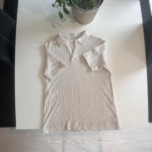 Beige medium pikétröja från Tiger of Sweden - Snygg beige pikétröja från Tiger of Sweden. Tröjan har klassisk krage, korta ärmar och är tillverkad i ett mjukt bomullstyg med struktur. Perfekt för en clean och stilren look eller för Old money stil eller grish stil.