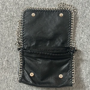 Svart axelväska med kedjedetaljer Tiamo - Säljer min superfina tiamo väska som inte längre kommer till användning! Den är äkta men har dock rostat lite på ringarna på sidorna som man kan se på bilden som håller ihop bandet till väskan och väskan. Skriv om ni har några frågor!💞ny pris är runt 500kr men priset kan diskuteras 