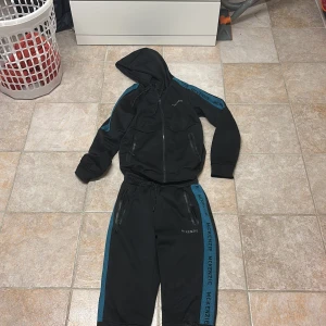 Svarta mjukisbyxor från McKenzie - Svarta mjukisbyxor från McKenzie med blå sidopaneler och logga längs benen. Byxorna har resår i midjan, snörning och dragkedjeförsedda fickor. Materialet är mjukt och stretchigt, perfekt för chill eller träning.