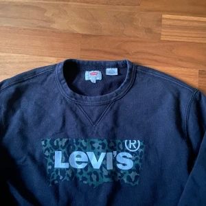 Svart Levi's sweatshirt med leopardmönster - Cool svart sweatshirt från Levi's med stort tryck framtill i blått och grönt leopardmönster. Klassisk rund halsringning och långa ärmar. Perfekt för dig som gillar streetstyle och vill sticka ut lite extra.