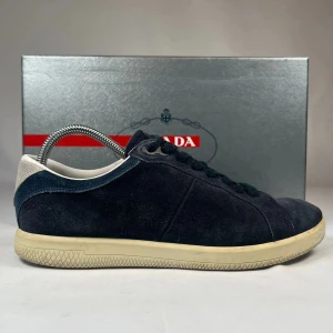 Mörkblå mockasneakers från Prada - Snygga mörkblå sneakers från Prada i mjuk mocka med beige sula och vit detalj vid hälen. Klassisk låg modell med rund tå och svarta snören. Diskret Prada-logga på plösen. Perfekta för dig som gillar stilrena och exklusiva sneakers.