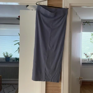 Lång blå kjol i slät polyester - En stilren långkjol i en dämpad blå/lila nyans. Kjolen har rak passform och är tillverkad i ett slätt polyestertyg som faller snyggt längs benen. Perfekt för dig som gillar minimalistisk och enkel stil.