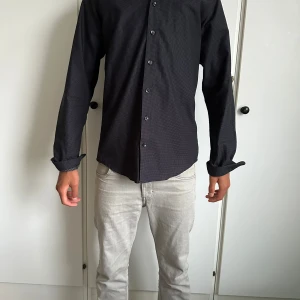 Svart slim fit skjorta Dressmann S - Svart skjorta från Dressmann i slim fit-modell, storlek S (37/38). Skjortan har ett diskret prickigt mönster, långa ärmar och knäppning framtill. Snygg och stilren passform som sitter tajt mot kroppen. Perfekt för dig som gillar en klassisk look med en twist.