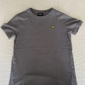  Mörk Grå t-shirt från Lyle & Scott - Snygg grå t-shirt från Lyle & Scott med klassisk rund hals och korta ärmar. På bröstet sitter den ikoniska gula fågel-loggan som broderad detalj. Perfekt basic-plagg i mjuk bomull som funkar till allt.