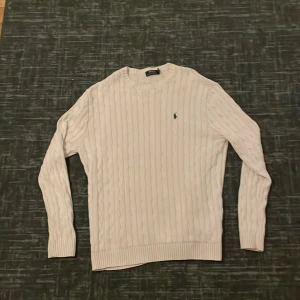 Vit kabelstickad tröja Polo Ralph Lauren - Kabelstickad vit tröja från Polo Ralph Lauren med klassisk rund hals och broderad logga på bröstet. Tröjan är långärmad och har ribbade muddar vid ärmslut och nederkant. Perfekt för en clean och stilren look.