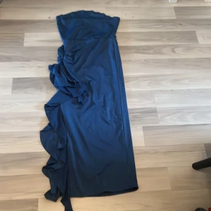 Marinblå långklänning med volang - Säljer en marinblå långklänning med snygga volangdetaljer längs sidan. Klänningen är axelbandslös och har en elegant, figurnära passform som ger en stilren look. Perfekt för dig som vill sticka ut med en klassisk men trendig stil.