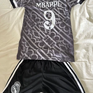 Real Madrid Mbappé fotbollströja Adidas - Snygg Real Madrid fotbollströja från Adidas med Mbappé och nummer 9 på ryggen. Tröjan är grå med ett unikt grafiskt mönster och vita detaljer, samt klubbmärke och sponsortryck framtill. Kortärmad modell i lätt och ventilerande material, perfekt för match eller träning. Man får även shorts till. Kan diskutera pris Skfic så fort ni är intresserade! Går med på de flesta😊