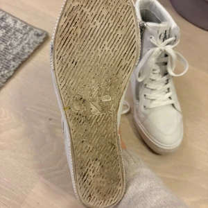 Vita höga sneakers från Calvin Klein - Snygga vita höga sneakers från Calvin Klein Jeans med stort CK-logo på sidan. Skorna har snörning, dragkedja på sidan och är gjorda i skinn. Platt sula och rund tå ger en clean look, perfekt för dig som gillar streetstyle med en twist. I mycket bra skick. 
