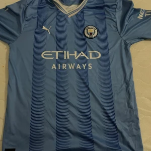 Manchester City Haaland matchtröja Puma - Manchester Citys officiella matchtröja i ljusblått med mörkblå ränder, Puma-logga och klubbmärke på bröstet. Framsidan har Etihad Airways-tryck, baksidan har HAALAND och nummer 9. Kortärmad och tillverkad i lätt funktionsmaterial.