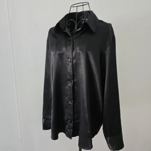 Svart satinliknande skjorta - Stilren svart skjorta i glansigt satinliknande material med klassisk krage och knäppning framtill. Skjortan har långa ärmar med manschett och en loose fit som ger en avslappnad men ändå dressad vibe. Det finns lite missfärgning på den inre delen av kragen, men det syns inte när skjortan används. I övrigt är skjortan i mycket gott skick.