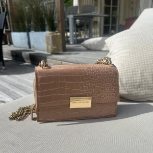 Beige axelväska från Ideal of Sweden - Snygg beige axelväska med krokopräglat mönster och guldfärgad kedja. Väskan har ett stilrent metallspänne framtill och flera fack inuti, inklusive ett med dragkedja. Perfekt för att bära det viktigaste med stil.