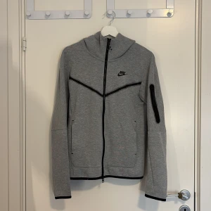 Grå Nike Tech Fleece set från Nike - Grå zip-hoodie från Nike med svart logga på bröstet och svarta detaljer. Jackan har hel dragkedja, huva och en ficka med dragkedja på ärmen, hoodien har storlek XS. Samt grå byxor som går ihop, byxorna har storlek S. Tillverkad i mjuk bomullsblandning, perfekt för chill eller träning.