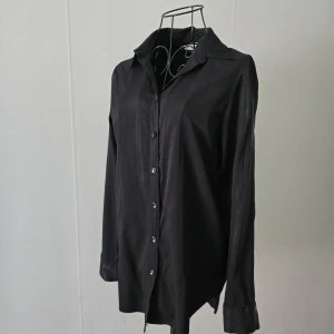Svart långärmad skjorta med knappar - En stilren svart skjorta med klassisk krage och knäppning framtill. Skjortan har långa ärmar med manschett och en rak, något loose passform. Baksidan av plagget har fått lite noppor, men i övrigt är det i gott skick