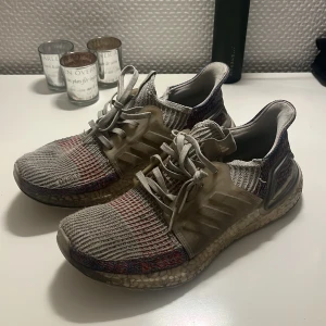 Adidas Ultraboost grå sneakers - Adidas Ultraboost sneakers i grått med stickad ovandel och inslag av rött och blått. Skorna har snörning, platt sula och en bekväm Boost-mellansula. Perfekta för dig som gillar sportig stil och vill ha maximal komfort i vardagen.