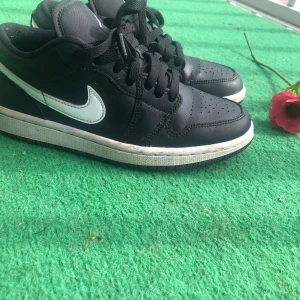 Svarta Nike Air Jordan 1 Low sneakers - Svarta Nike Air Jordan 1 Low med vit swoosh och vit sula. Klassisk siluett med perforerad tå, snörning och låg profil. Skorna är i skinn och har en stilren look som passar till streetwear. Perfekta för dig som gillar sneakers med ikonisk design.