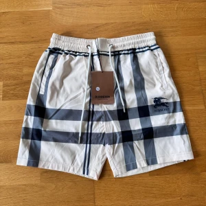 Burberry rutiga badshorts med snörning - Snygga badshorts från Burberry i klassiskt rutigt mönster i vitt och mörkblått. Shortsen har elastisk midja med snörning och Burberry-logga på ena benet. Perfekta för sommaren och strandhäng, tillverkade i lätt och snabbtorkande material.