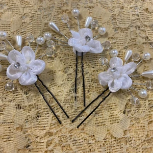 Vita blommiga hårnålar med pärlor - Tre hårnålar dekorerade med vita tygblommor, glittrande stenar och pärlliknande detaljer. Perfekt för att ge en romantisk och elegant touch till frisyren. Blommorna har en kristall i mitten och omges av pärlor och transparenta pärlor på silverfärgade trådar.