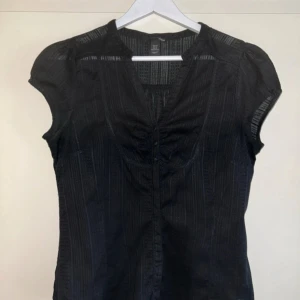 Svart blus med knappar från H&M - Svart blus från H&M med subtila ränder i tyget, v-ringning och små knappar framtill. Tunt, lätt material och korta ärmar med lätt puff. Perfekt för dig som gillar en stilren look med lite extra detalj. Storlek:XL men skulle säga M/L 