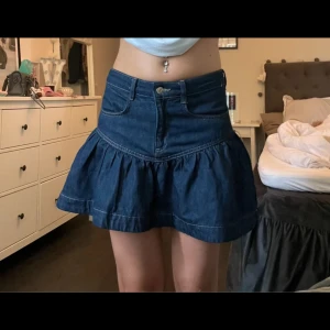 Mörkblå jeanskjol med volang - Supersöt mörkblå jeanskjol med volang och mid waist från H&M. Men den går att vika ner om man vill ha den low waist. Kjolen har klassiska jeansfickor fram och bälteshällor. Stängs med knapp och dragkedja. Perfekt för dig som gillar en trendig och avslappnad stil. Har använt den 1-2 gånger så fortfarande i ett nytt skick. 