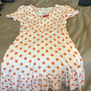 Vit kortklänning med orange prickar - Superfin vit kortklänning från LCW Casual med stora orange prickar, puffärmar och knappar framtill. Klänningen har volang nertill och en söt, feminin passform som känns både lekfull och trendig. Perfekt för dig som gillar färg och mönster!