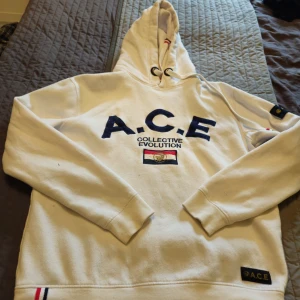 Vit hoodie från A.C.E Collective Evolution XL - Vit hoodie från A.C.E Collective Evolution med broderad logga och text i mörkblått på bröstet. Tröjan har huva med snörning, ribbade muddar och detaljer i rött, vitt och blått. Snygg patch på ärmen och nertill. Perfekt för dig som gillar streetwear. Liten i storlek