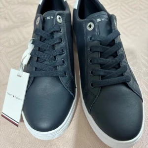 Svarta sneakers från Tommy Hilfiger - Snygga svarta sneakers från Tommy Hilfiger med vit logga på sidan och vit häl. Skorna har snörning, rund tå och platt vit sula. Tillverkade i skinn för en clean och stilren look. Perfekta för dig som gillar klassisk streetstyle.