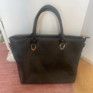 Svart axelväska i skinnimitation - Stilren svart axelväska med både handtag och lång axelrem. Den har fina gulddetaljer och blommigt mönster i beige och svart. Perfekt för dig som vill ha plats för allt du behöver under dagen. Jag köpte den secondhand men har alldrig användt den för jag har för många väskor