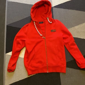 Röd hoodie från Henri Lloyd - Riktigt snygg röd hoodie från Henri Lloyd med dragkedja och huva. Tröjan har vita snören med detaljer, två stora fickor framtill och ett litet märke med brittisk flagga på bröstet. Perfekt för dig som gillar klassisk streetstyle och vill sticka ut.