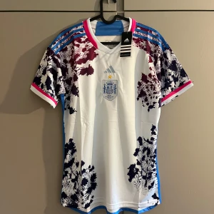 Spanien bortatröja Adidas 2024 - Säljer Spaniens bortatröja från Adidas med vit bas, blå och vinröda detaljer samt unikt mönster på axlar och ärmar. Tröjan har korta ärmar, blå Adidas-ränder och broderat landslagsmärke på bröstet. Tillverkad i lätt och ventilerande material.