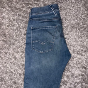 Replay Anbass ljusblå slim jeans - Snygga ljusblå jeans från Replay, modell Anbass. Slim fit med klassisk femficksdesign och diskreta slitningar. Jeansen är tillverkade i stretchigt Hyperflex-material för extra komfort och har en modern, smal passform. Perfekta för en avslappnad och trendig look.