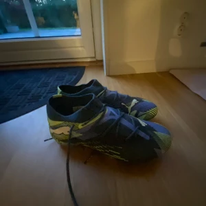 Puma fotbollsskor i blått och grönt  - Säljer ett par snygga Puma fotbollsskor med coolt mönster i blått, svart, grönt med vita ditaljer. Skorna har snörning och är tillverkade i syntetmaterial med mönstrad ovandel och dobbar för bra grepp på planen, är till både gräs och konstgräs. HAR INTE ANVÄNDS LÄNGE!! Säljer pågrund av lite för liten storlek