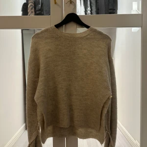 Beige stickad tröja med snörning - Mysig beige stickad tröja med lång ärm och rund hals. Tröjan har en loose passform och snörningsdetalj vid ärmsluten. Perfekt för lager-på-lager och chill höst- eller vinterstil.