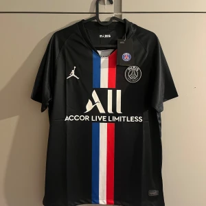 PSG x Jordan svart matchtröja - Svart PSG fotbollströja i samarbete med Jordan. Tröjan har korta ärmar, klassisk passform och är gjord i lätt, ventilerande material. Framtill finns vertikala blå, vita och röda ränder, PSG-logga och Jordan-logga samt sponsortryck.