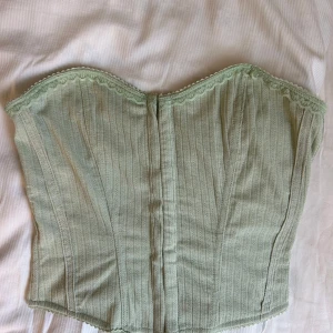 Ljusgrön korsett med spetskant - Säljer en ljusgrön korsett med fina spetsdetaljer längs överkanten. Toppen har en subtilt randig struktur och stängs med hyskor i ryggen. Perfekt för dig som vill ha en trendig och feminin look med vintagekänsla.