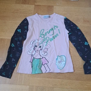 Rosa långärmad tröja  - Söt rosa långärmad tröja från Check This by Lindex Girls med svarta ärmar dekorerade med färgglada rosetter. Framsidan har ett tryck med texten 'Going to Paris!' och en tecknad figur. Tröjan är i mjuk bomull och har v-ringning. Är barnstorlek 145/152 men passar xs, eventuellt s. 🩷