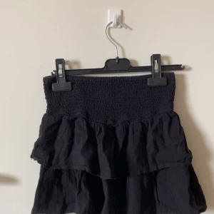 Svart volangkjol med smock - Supersöt svart kortkjol med bred smock i midjan och dubbla volanger. Kjolens lapp är bort klippt men den passar mig som vanligtvis har storlek S men skulle också säga att den passar Xs-M då den är stretchig