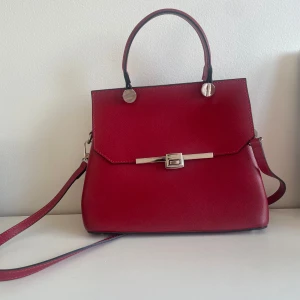 Röd handväska med silverdetaljer - Snygg röd handväska i skinnimitation med stilrena silverdetaljer och lås framtill. Väskan har både handtag och axelrem, samt praktiskt fack med dragkedja inuti. Perfekt för dig som vill ha en klassisk men trendig accessoar. Använd 1 gång