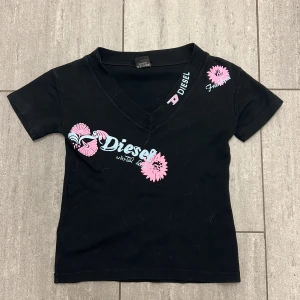 Svart t-shirt med tryck från Diesel - Svart t-shirt från Diesel med v-ringning och korta ärmar. Framsidan har coolt tryck i rosa och ljusblått med blommor och Diesel-logga. Materialet är 100% bomull, vilket gör den skön och lätt att bära. Passformen är normal och t-shirten har en ungdomlig vibe.