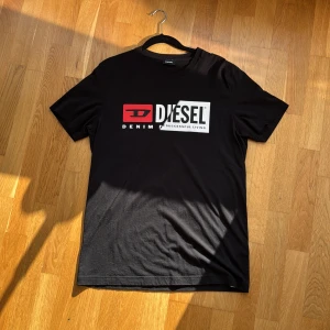 Svart t-shirt från Diesel med logga - Svart t-shirt från Diesel med stor vit och röd logga framtill. Klassisk rund halsringning och korta ärmar. Tillverkad i mjuk bomull som känns skön mot huden. Perfekt för dig som gillar streetwear och vill ha en enkel men snygg look. Snygg detalj som går diagonalt över framsidan på t-shirten. 