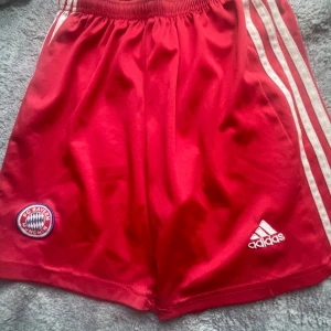Röda FC Bayern München shorts Adidas - Snygga röda fotbollsshorts från Adidas med FC Bayern München-logga på ena benet och klassiska vita ränder längs sidorna. Elastisk midja för skön passform. Perfekta för fotbollsträning eller när du vill köra sportig stil.