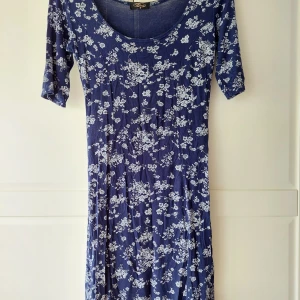 Blå blommig klänning - En mörkblå klänning med blommigt mönster i vitt/grått. Klänningen har en rund halsringning och ärmlängd till armvecket. Den är knälång och har en normal passform. Strechig sval och skön.