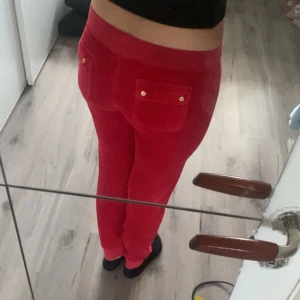 Röda Juicy Couture mjukisbyxor S - Riktigt snygga röda mjukisbyxor från Juicy Couture med guldiga detaljer och broderad logga. Byxorna har snörning i midjan, bakfickor med knappar och är gjorda i mjuk skön velour. Perfekta för en chill och trendig look.