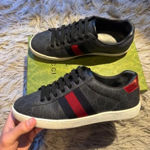 Gucci ace skor - Snygga svarta Gucci sneakers med klassiska röda och blå ränder på sidan och diskret GG-mönster i tyget. Skorna har svarta skosnören, vit platt sula och en röd metallic-detalj på hälen. 7/10 skick bara för att sulan är lite smutsig och de är en liten tråd på sidan av skon som låsstnat lite men inget som syns. Bara att skriva om ni vill se fler bilder på defekterna. Allt OG kommer med skorna. Bara att skriva vid minsta fundering!🖤❤️