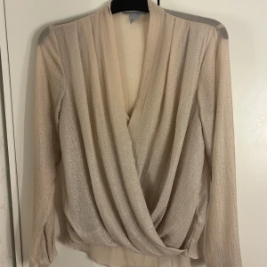 Glittrig omlottopp  - Säljer en superfin glittrig omlottopp från H&M i beige med lång ärm. Toppen har en djup v-ringning och ett snyggt omlottfall framtill. Materialet är lätt och skimrande. Vid frågor är det bara att höra av sig!💞