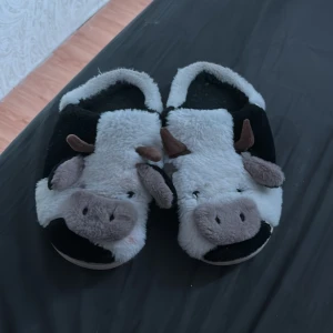 Fluffiga tofflor med ko-design - Supermysiga tofflor i svart och vitt med gulligt ko-ansikte, öron och horn framtill. Tillverkade i mjuk syntetpäls och med platt sula. Perfekta för att chilla hemma och hålla fötterna varma under kyliga dagar.