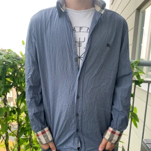 Blå randig skjorta från Burberry - Snygg blå skjorta från Burberry med tunna vita ränder och klassiska rutiga detaljer på insidan av krage och ärmslut. Skjortan har knappar framtill och lång ärm, samt diskret Burberry-logga broderad på bröstet. Perfekt för dig som gillar stilrena plagg med en twist.